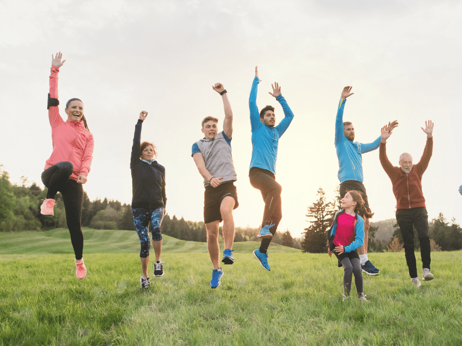 Gruppe von acht Personen springt fröhlich auf einer Wiese vor einem bewaldeten Hintergrund in der Abenddämmerung.