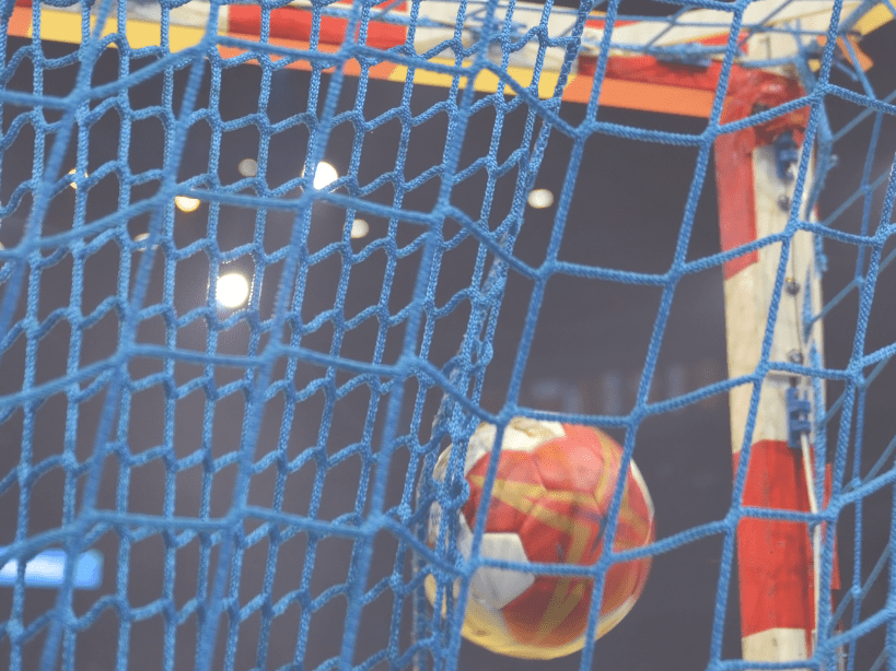 Nahaufnahme eines Handballs, der ins Netz eines Torrahmens aus blauen und weißen Schnüren fliegt.