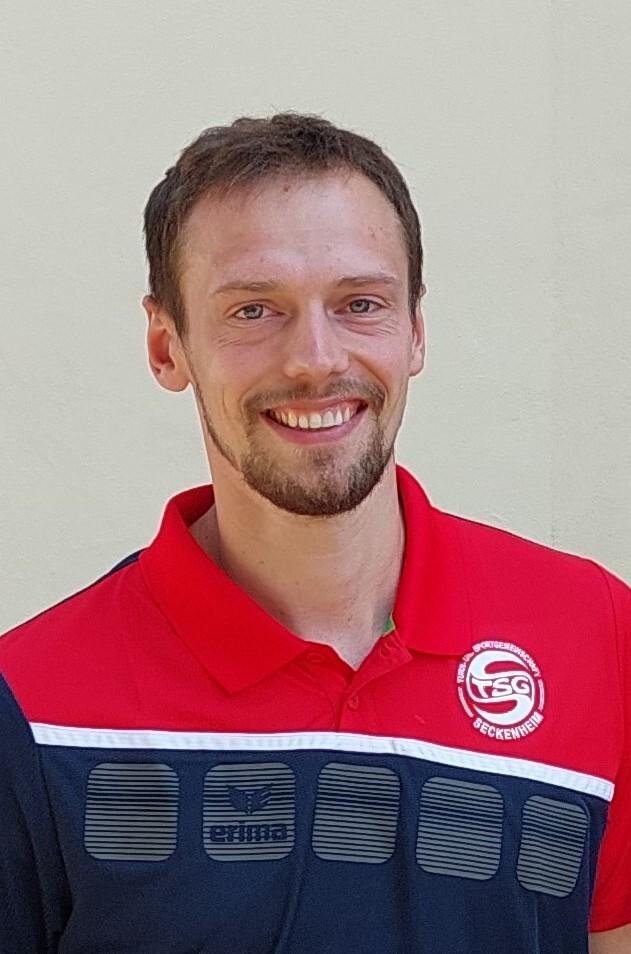 Lächelnder Mann mit Kurzhaarschnitt trägt ein rotes Sportshirt mit TSG-Logo vor einer einfarbigen Wand.