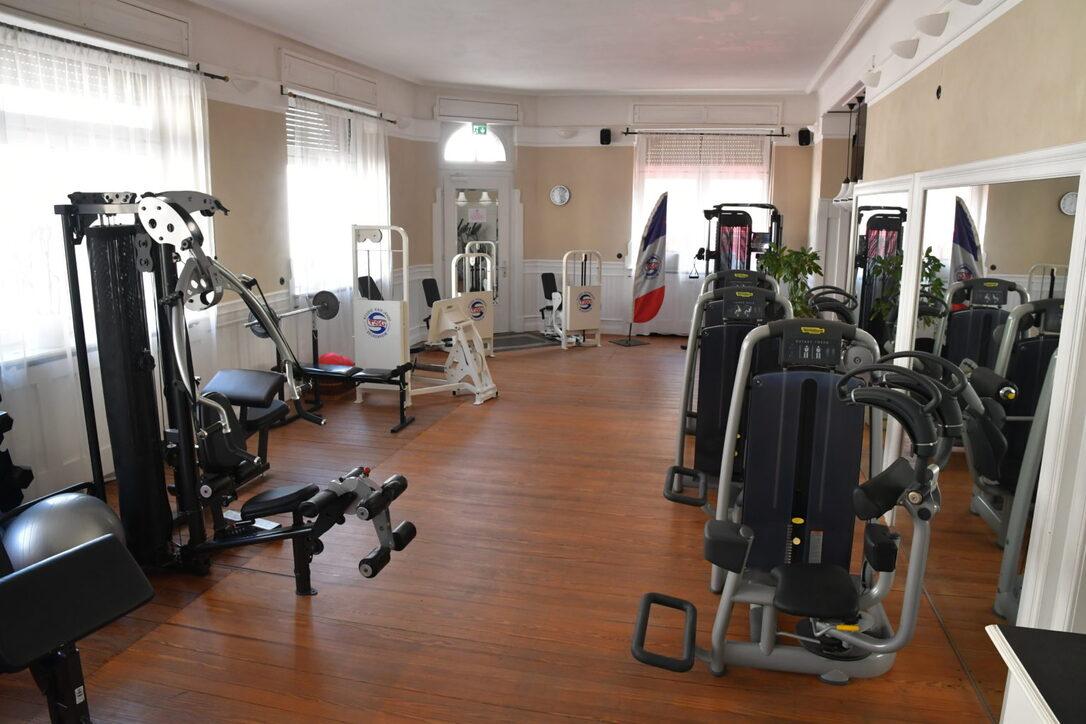 Fitnessraum mit verschiedenen Kraftgeräten, hölzernem Boden und großen Fenstern, die Tageslicht hereinlassen.