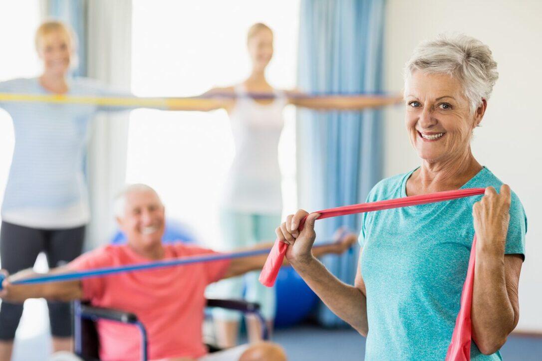 Ältere Frau hält ein rotes Widerstandsband, während andere im Hintergrund mit elastischen Bändern trainieren.