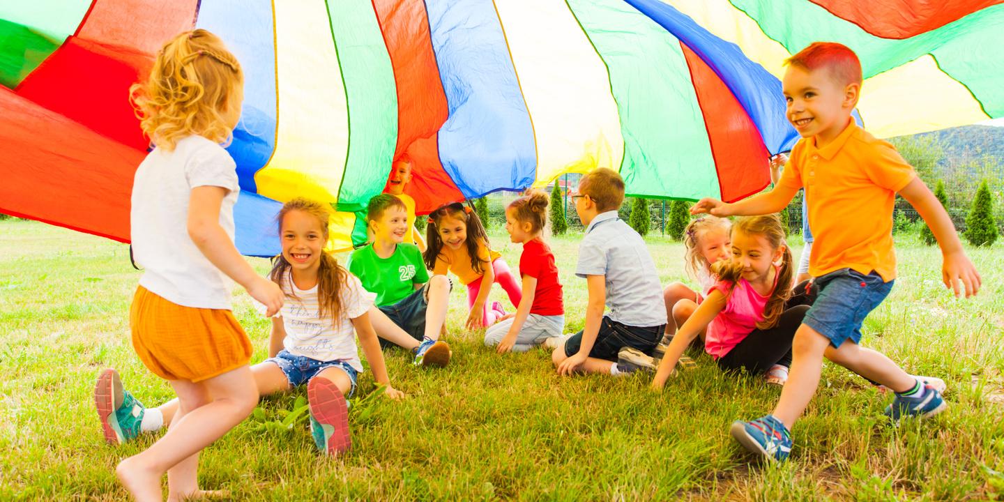 Eine Gruppe von Kindern sitzt unter einem bunten Spieltuch auf der Wiese und lacht miteinander.