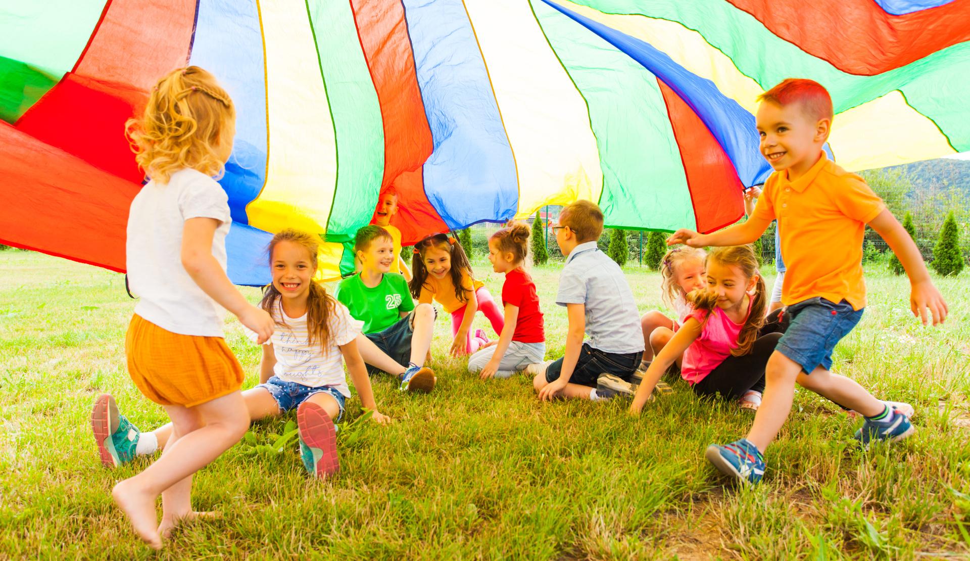Eine Gruppe von Kindern spielt unter einem bunten Tuch auf einer Wiese und hat Spaß miteinander.