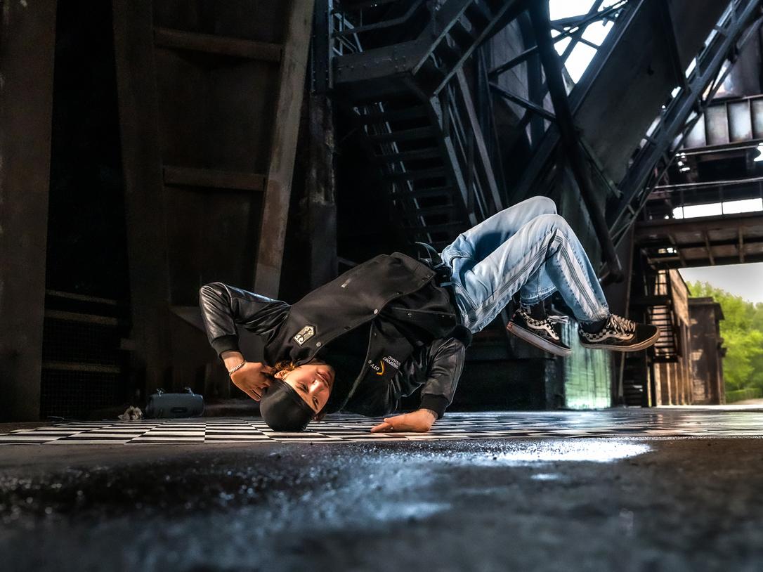Breakdancer in schwarz-beige Kleidung führt eine Kopfstandbewegung auf einem fliesenartigen Boden in einer Industrieumgebung aus.
