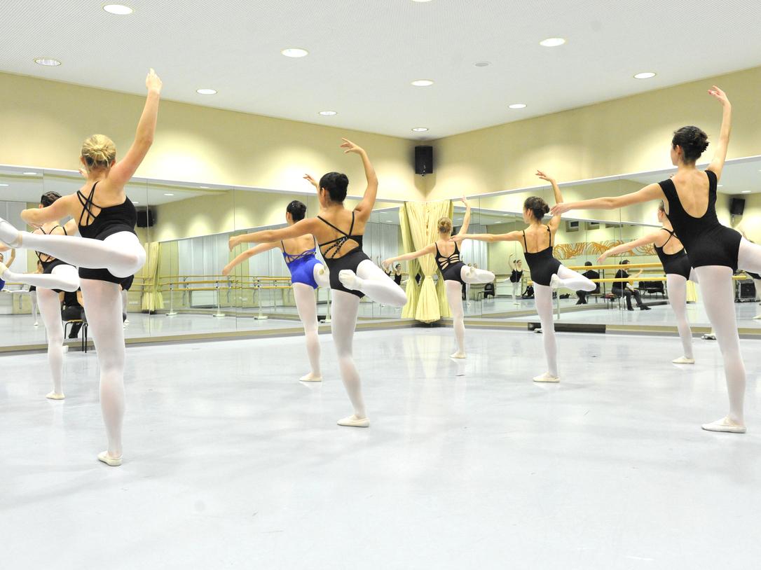 Gruppe von Balletttänzerinnen in Schwarz und Blau übt in einem Spiegelraum mit Holzballettstangen.