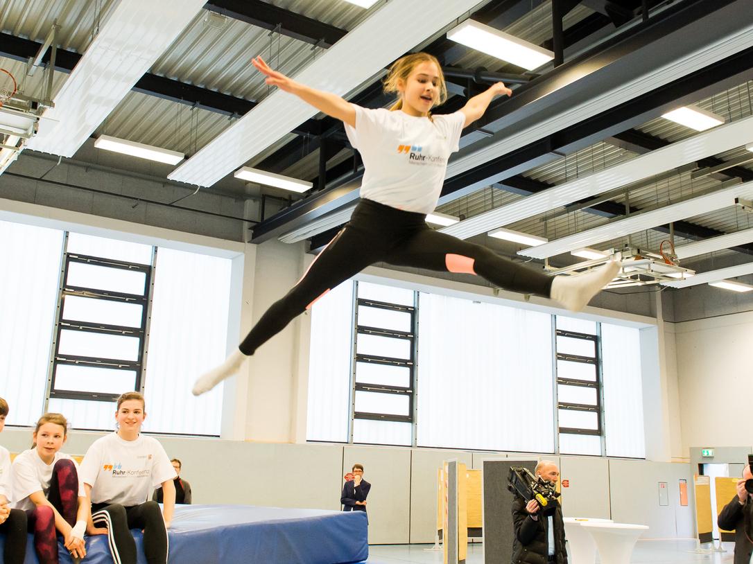 Mädchen springt mit ausgestreckten Armen über eine Trampolinfläche, während Zuschauer auf einer Bank sitzen.